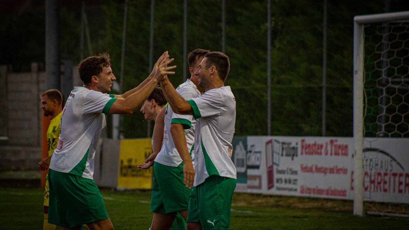 Drei Fußballspieler in grünen und weißen Trikots feiern auf einem Feld bei Nacht mit einem verschwommenen Hintergrund.