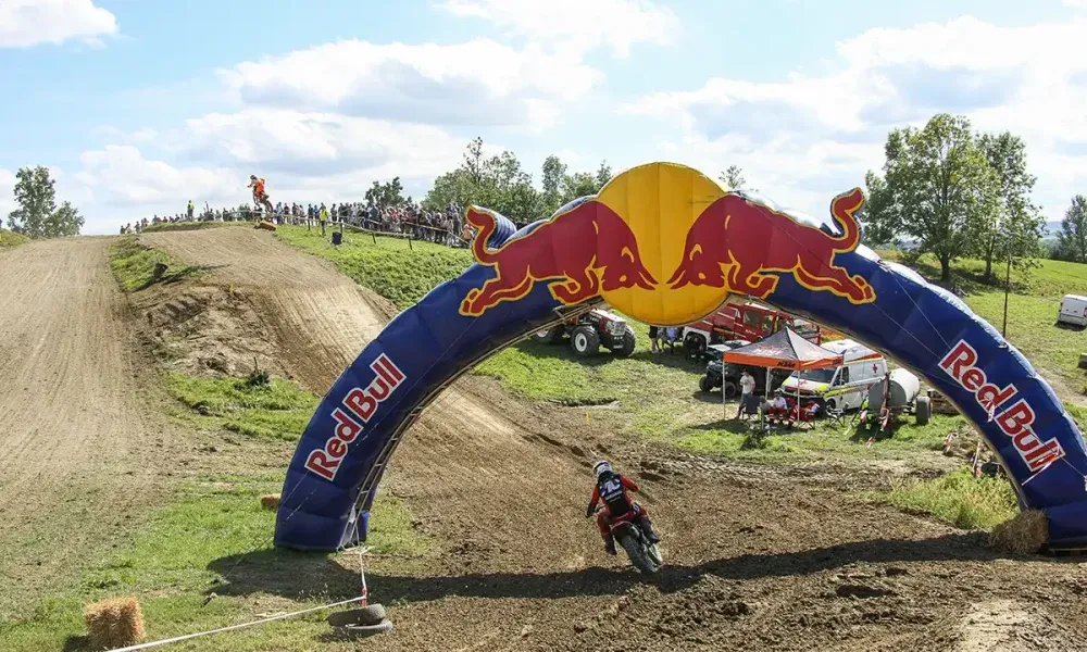 A motocross rider rides under a Red Bull arch on a dirt track with a crowd watching.