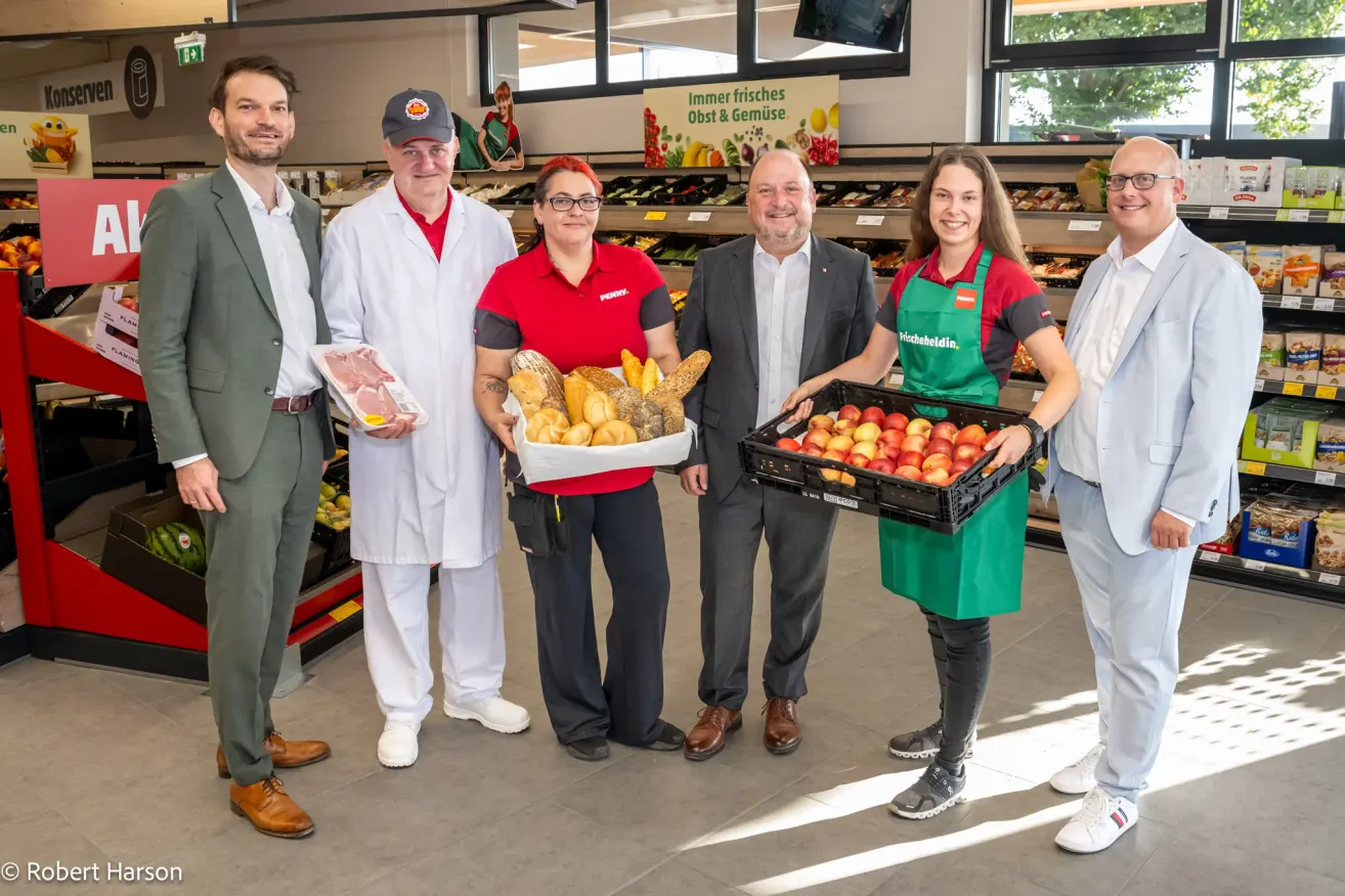 Eine Gruppe von Menschen in einem Supermarkt, zwei Frauen halten Körbe mit Brot und Obst, ein Mann hält ein Paket mit Fleisch und zwei Männer in Anzügen lächeln.