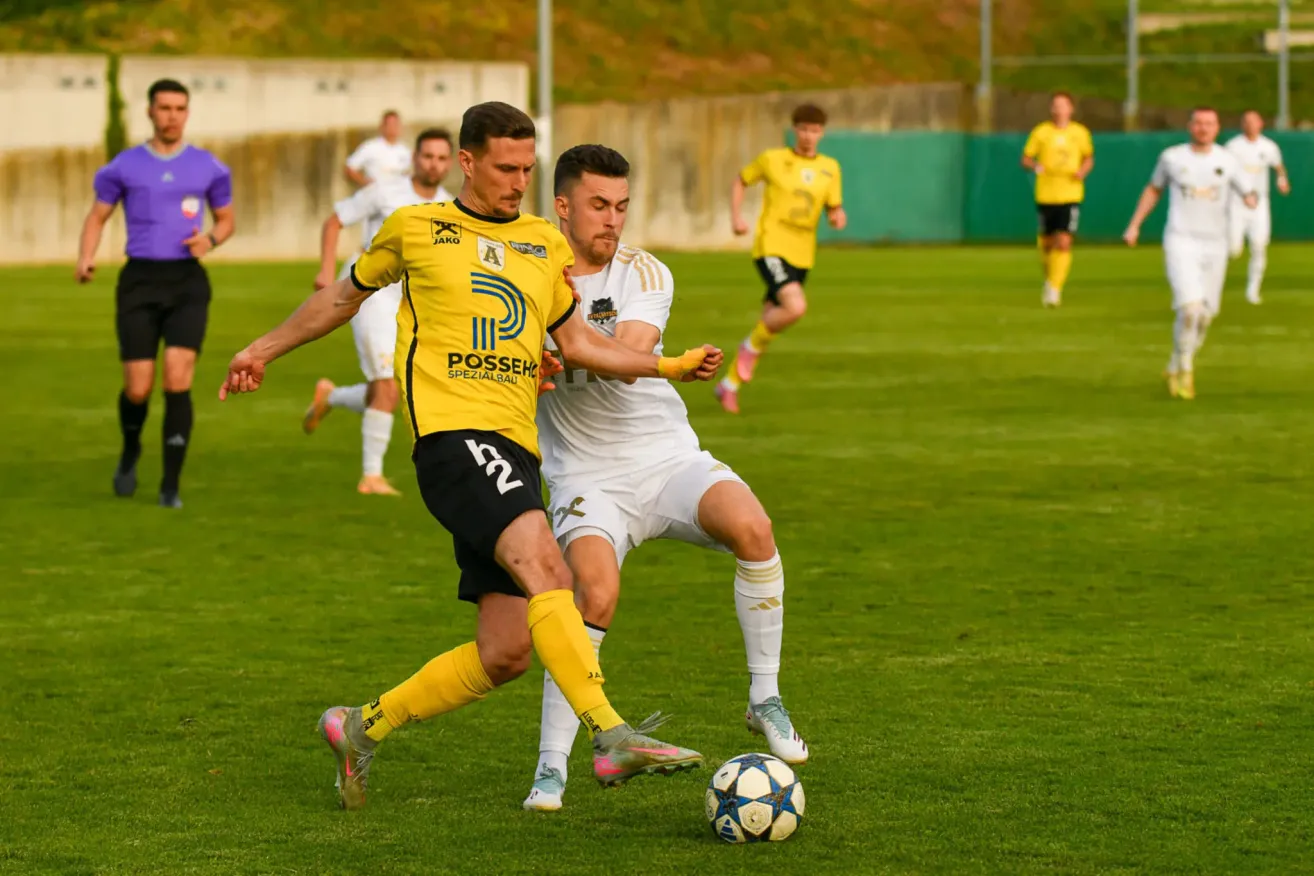 Zwei Fußballspieler in gelben und weißen Trikots sind in einem dynamischen Duell um den Ball auf einem grünen Feld. Der Spieler in Gelb versucht, dem Spieler in Weiß den Ball zu stehlen.