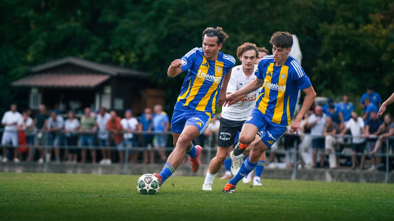 Drei Fußballspieler in blau-gelben Trikots laufen auf dem Feld. Ein Spieler tritt den Ball, während die anderen ihn verfolgen. Zuschauer beobachten von den Rändern.