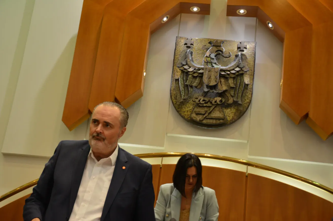 Ein Mann in einem Anzug steht neben einer Frau in einem Blazer, beide sehen ernst aus. Hinter ihnen ist eine Wand mit einem goldenen Emblem.