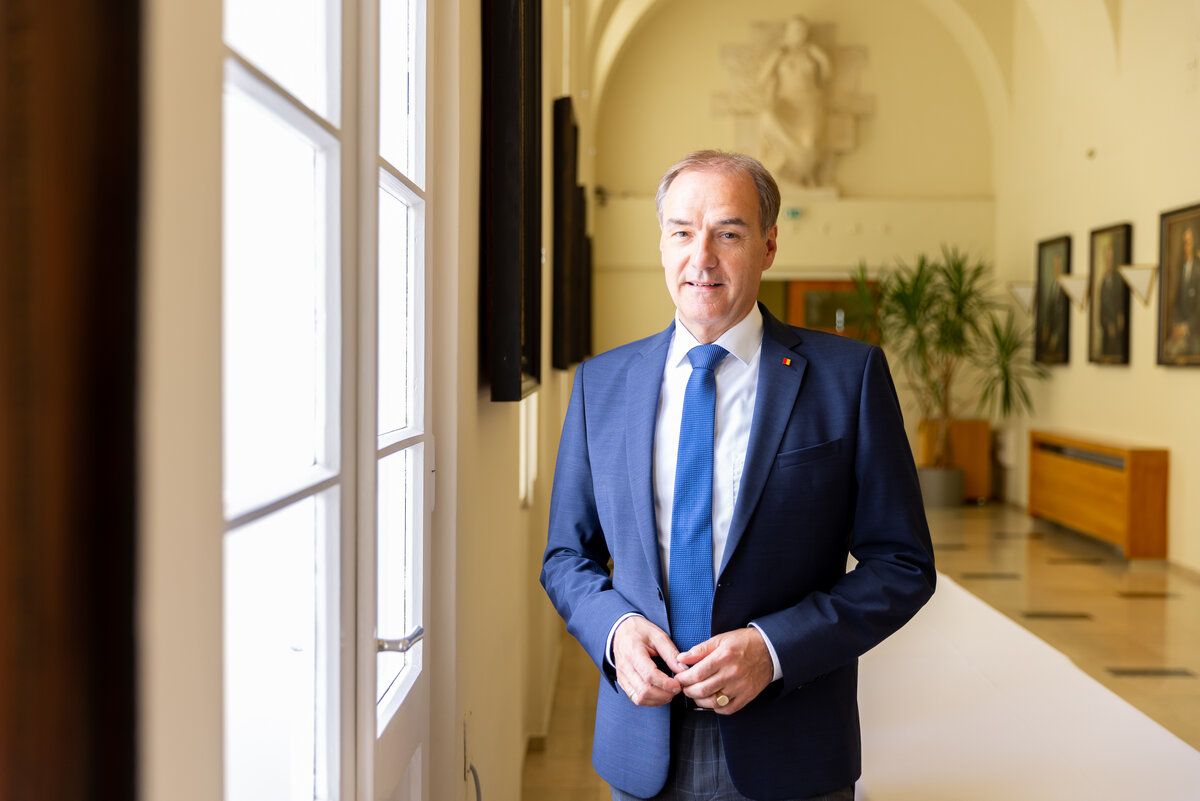 A man in a blue suit stands in a hallway with arched ceilings, smiling and looking at the camera. Behind him, there is a glass door and a potted plant.