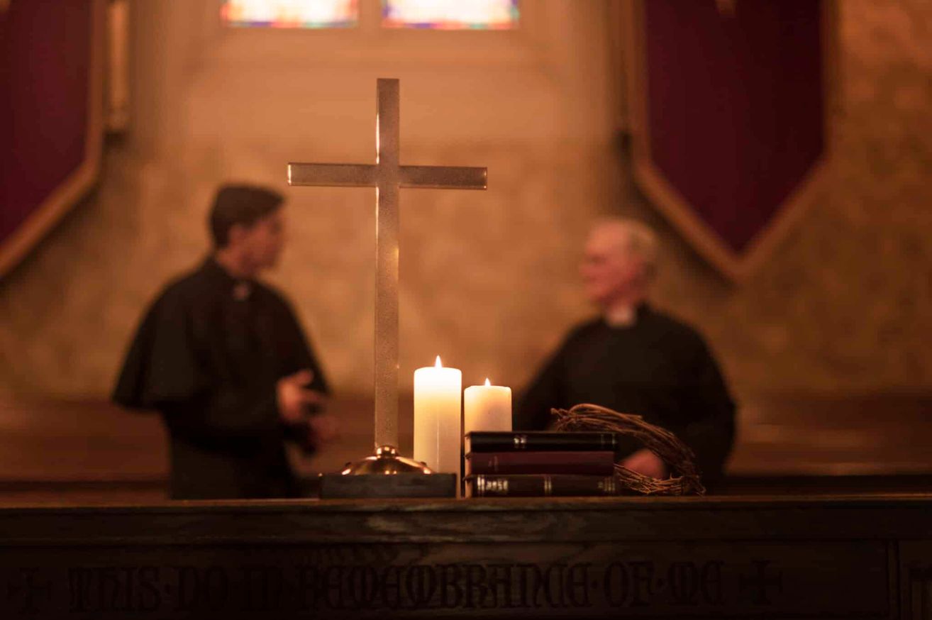 Zwei Priester stehen hinter einem Tisch mit einem Kreuz, Kerzen und Büchern in einer schwach beleuchteten Kirche.