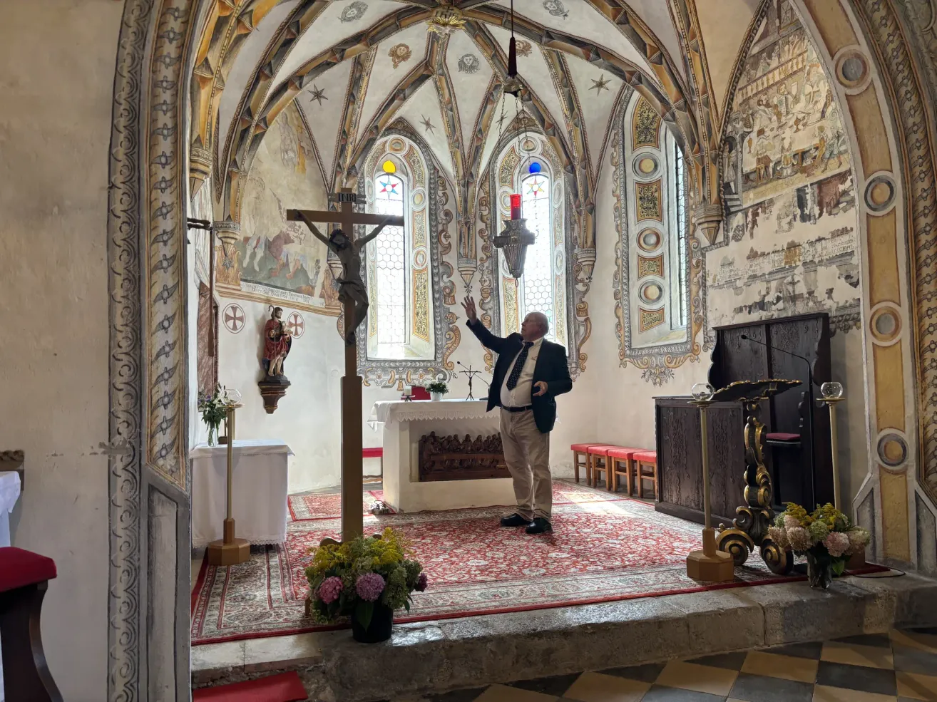 Ein Mann in Anzug steht in einer Kirche und zeigt auf ein Kruzifix auf einem Podest vor einem geschmückten Altar.