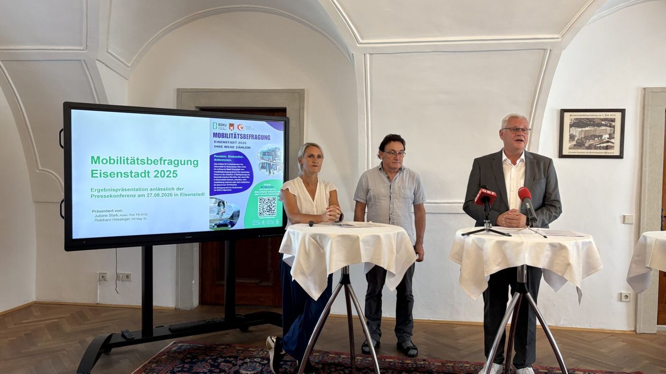 Drei Personen stehen hinter einem Tisch mit Mikrofonen, einer von ihnen spricht, während eine große Leinwand eine Präsentation über Mobilität im Jahr 2025 zeigt.