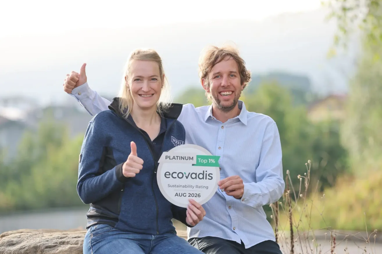 A man and woman are sitting on a rock, smiling and giving a thumbs up. The woman is holding a round sign that reads Platinum ecovadis Sustainability Rating Aug 2026. They are both wearing casual clothing. Behind them is a blurry background of trees and mountains.