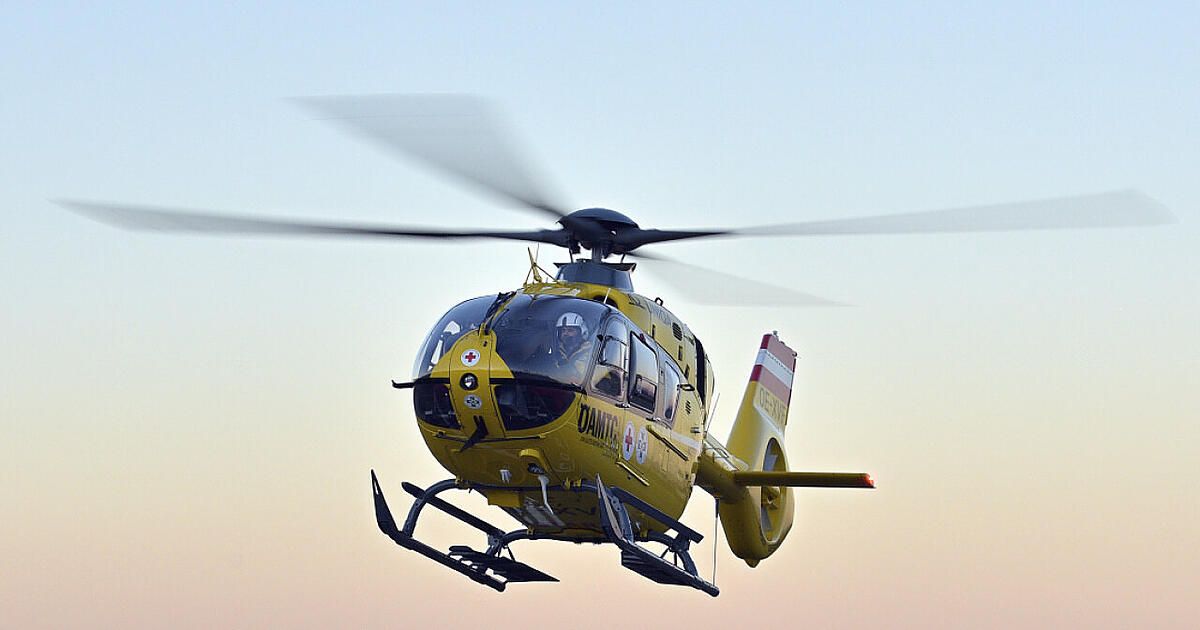 A yellow helicopter with red cross symbols on the side and a red tail is flying in the sky. The pilot is wearing a helmet.