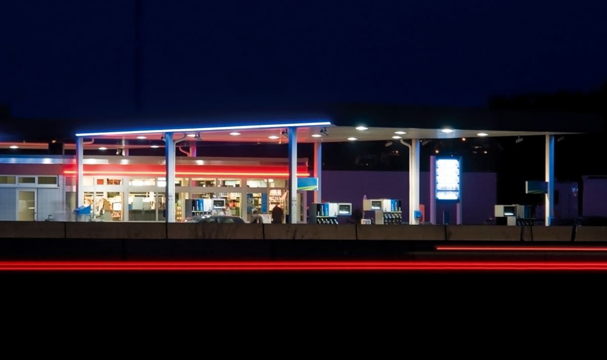 Eine Tankstelle bei Nacht mit roten und blauen Lichtern, die den Bereich beleuchten. Zwei Personen stehen in der Nähe der Zapfsäulen.
