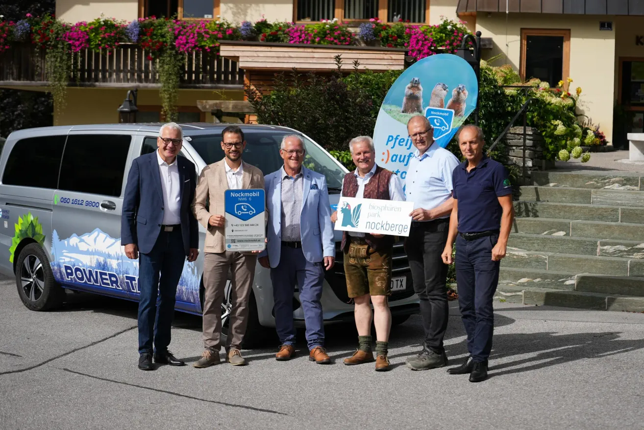 Five men stand in front of a van with a sign that reads 'Pfeil auf' and another sign that says 'Nockmobil NMG 6'. The man in the middle holds a certificate. Behind them, a building with a balcony adorned with flowers.
