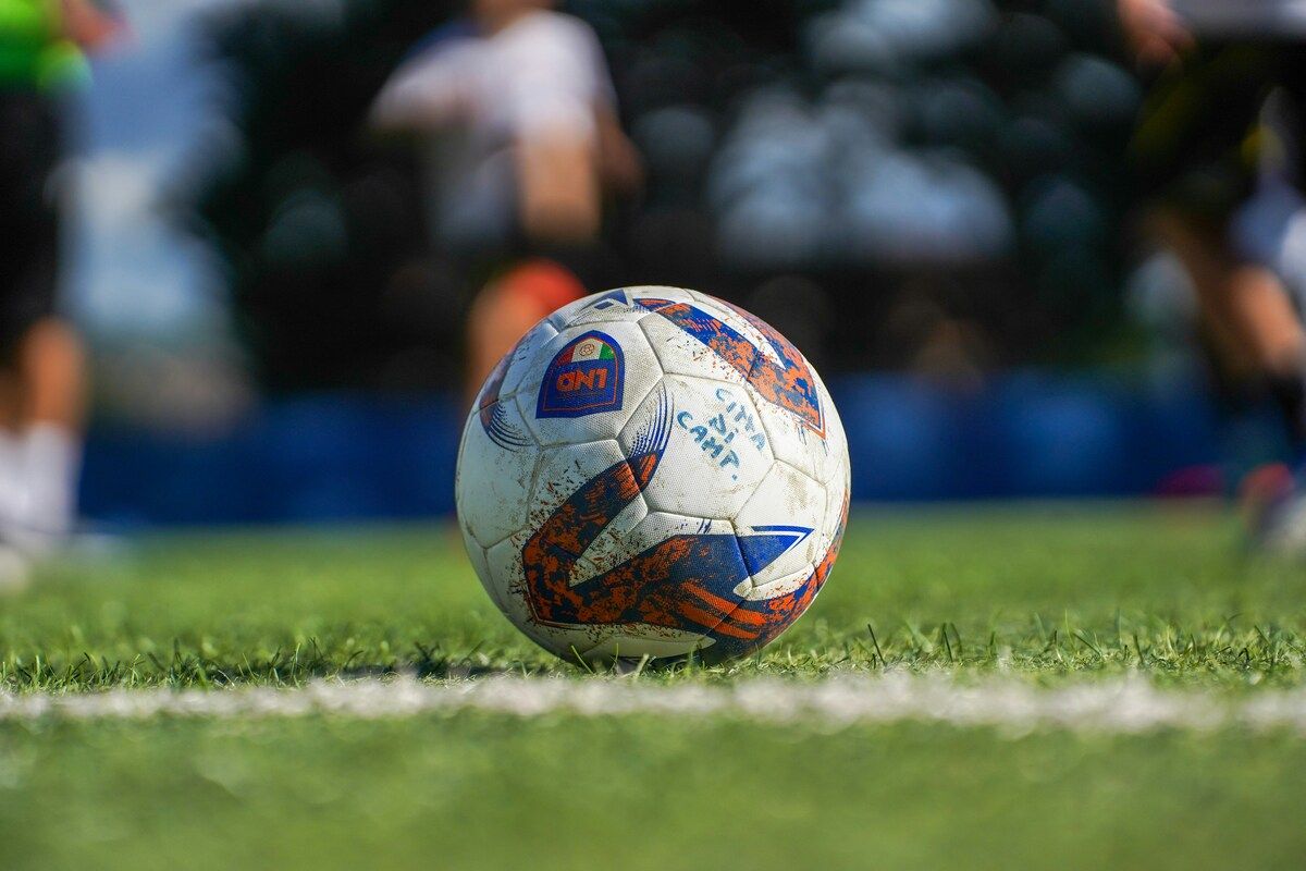 A soccer ball with a blue and orange design sits on a grassy field with a white line. In the background, a blurry figure of a person is visible.