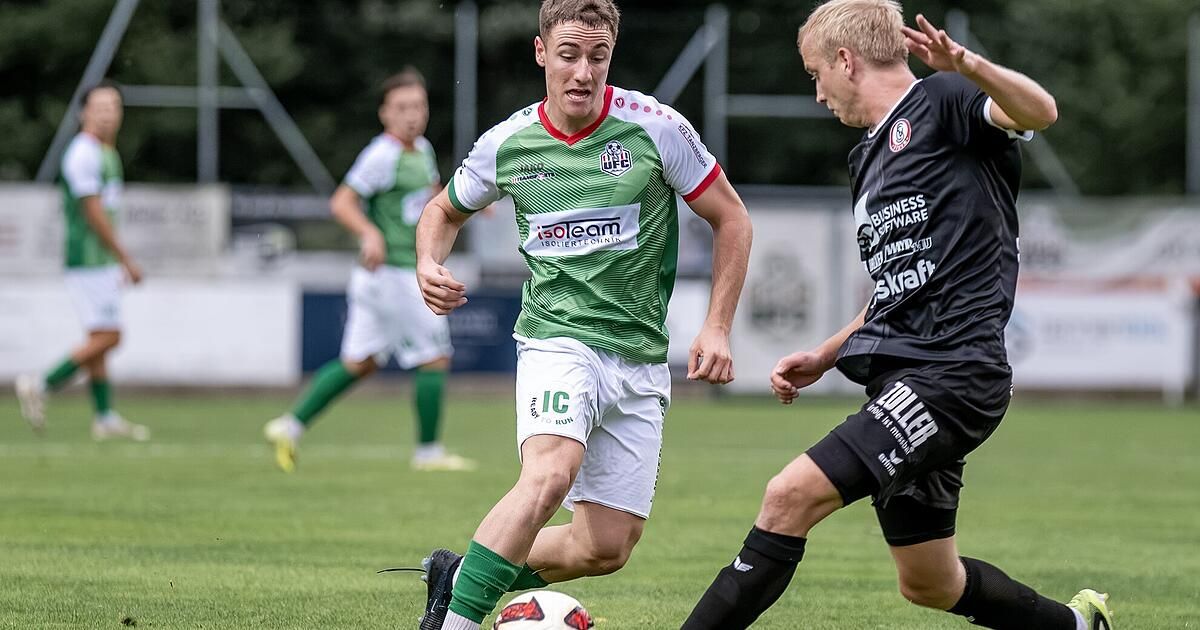 Ein Fußballspieler in einem grün-weißen Trikot läuft mit dem Ball, während ein anderer Spieler in Schwarz versucht, ihn zu blockieren. Im Hintergrund beobachtet ein weiterer Spieler in Grün und Weiß.