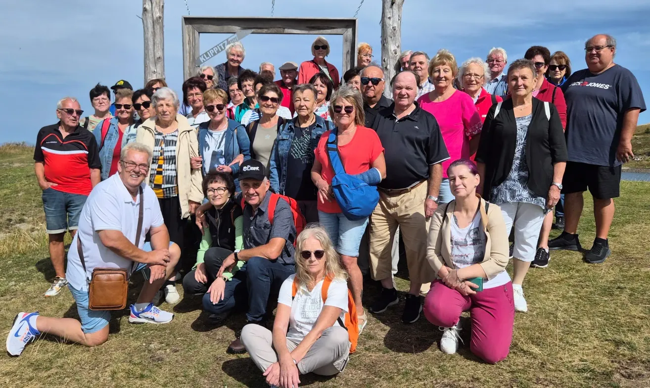Eine Gruppe von Menschen posiert für ein Foto im Freien, einige stehend und einige sitzend, mit einem Holzschild im Hintergrund.
