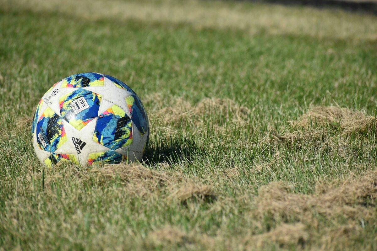 Ein Fußball mit einem mehrfarbigen Design, einschließlich Blau, Gelb und Rot, liegt auf einem Grasfeld. Der Ball zeigt das Adidas-Logo und das FIFA-Logo.