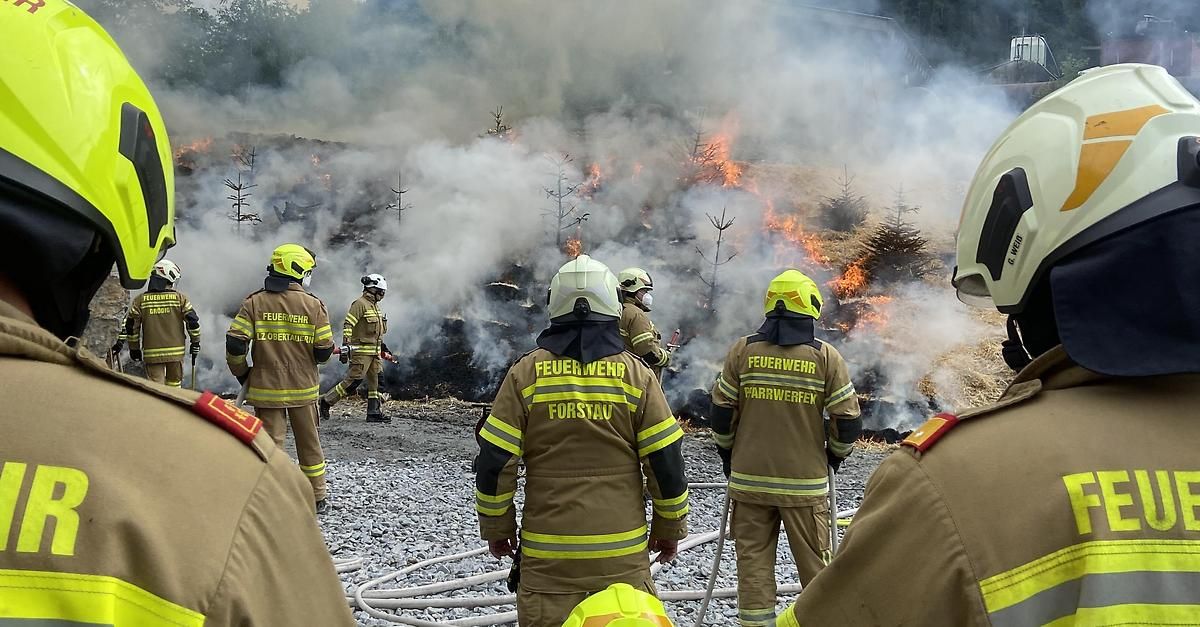 Feuerwehrleute in Schutzkleidung stehen in der Nähe eines brennenden Waldgebiets, einer hält einen Schlauch und ein anderer geht auf das Feuer zu.
