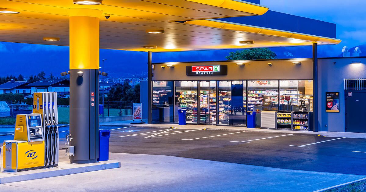 Eine Spar Express Tankstelle bei Nacht mit gelben Lichtern, die den Bereich beleuchten, ein klarer Blick in das Geschäftinnen mit Regalen, die mit Produkten gefüllt sind, und ein klarer Nachthimmel im Hintergrund.