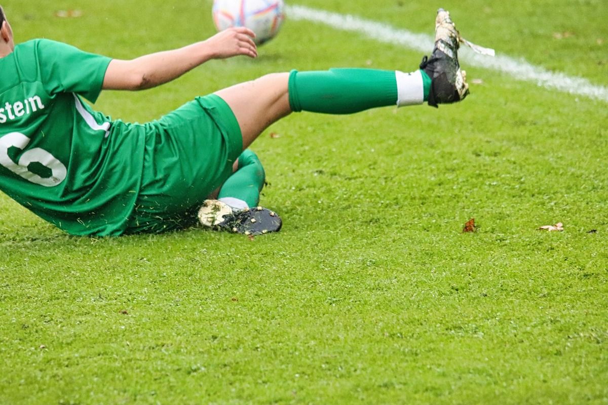 Ein Fußballspieler in einem grünen Trikot liegt auf dem Rasen, möglicherweise nach einem Sturz, mit einem Fußball in der Nähe.