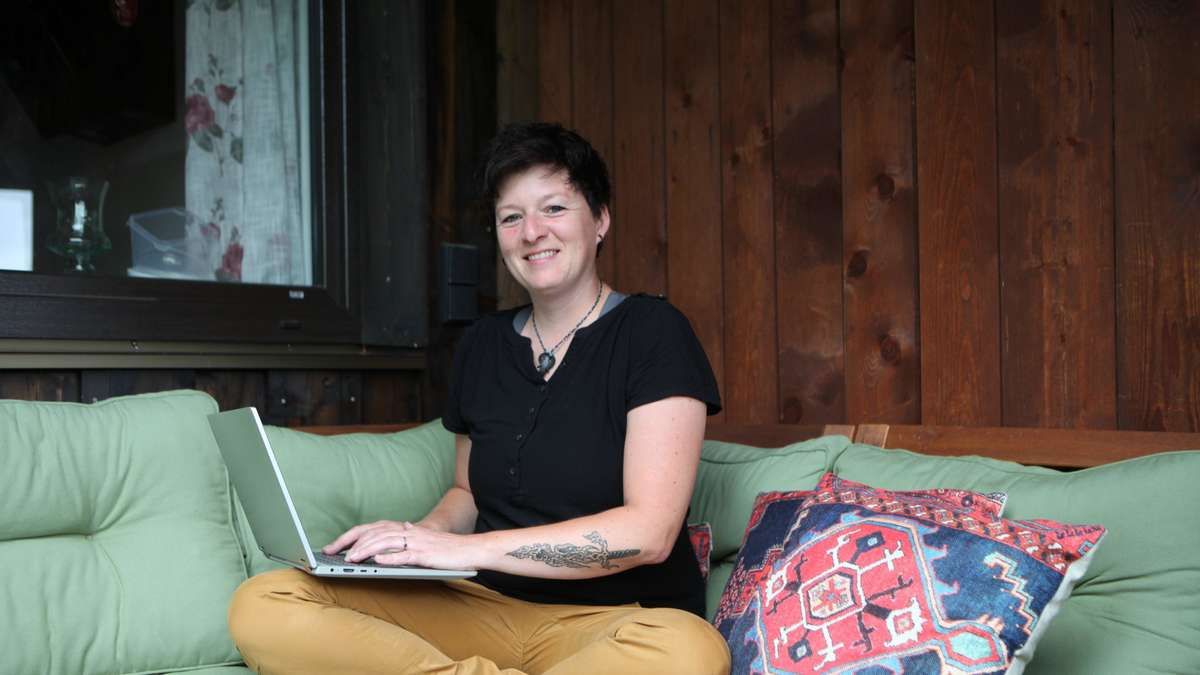 Eine Frau mit kurzen Haaren und einem Tattoo auf dem linken Arm sitzt auf einem Sofa, lächelt und benutzt einen Laptop. Hinter ihr befindet sich eine Holzwand mit einem Fenster.
