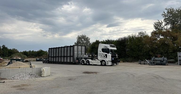 A large white semi-truck with a large black container attached is parked in a gravel lot with trees and bushes in the background.