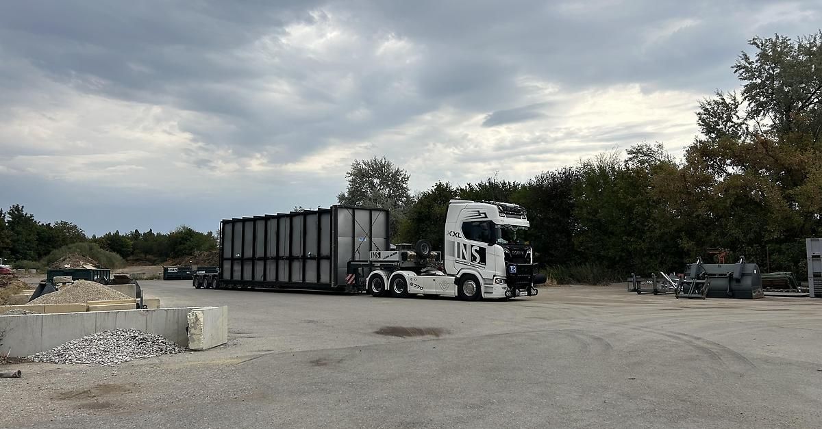 A large white semi-truck with a large black container attached is parked in a gravel lot with trees and bushes in the background.