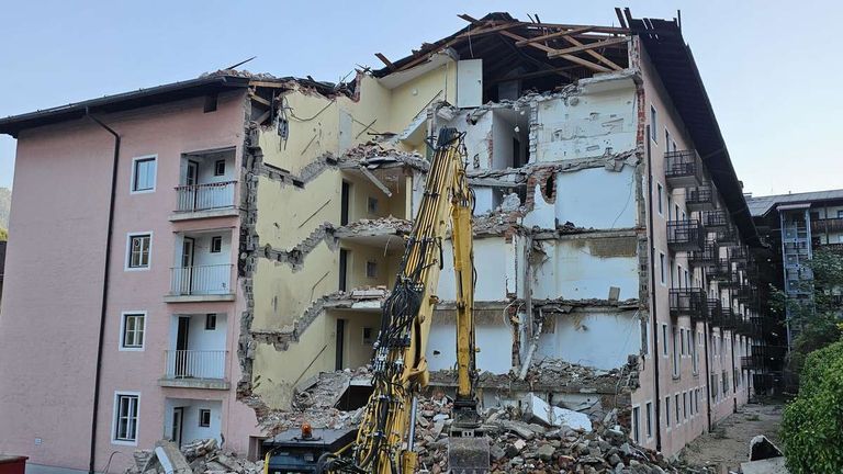 A yellow excavator is demolishing a multi-story building with a damaged roof and exposed interior.