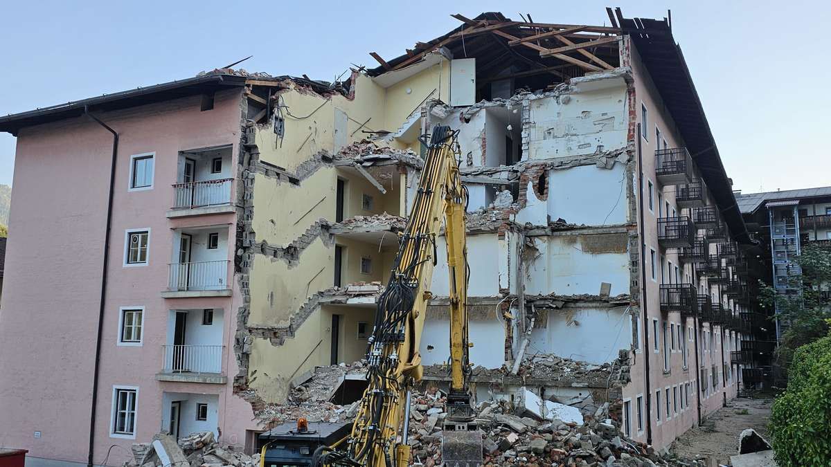 A yellow excavator is demolishing a multi-story building with a damaged roof and exposed interior.