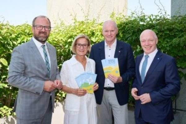 Four people dressed in formal attire are standing together in front of a green bush, smiling for a photo. The woman in the middle holds a booklet.