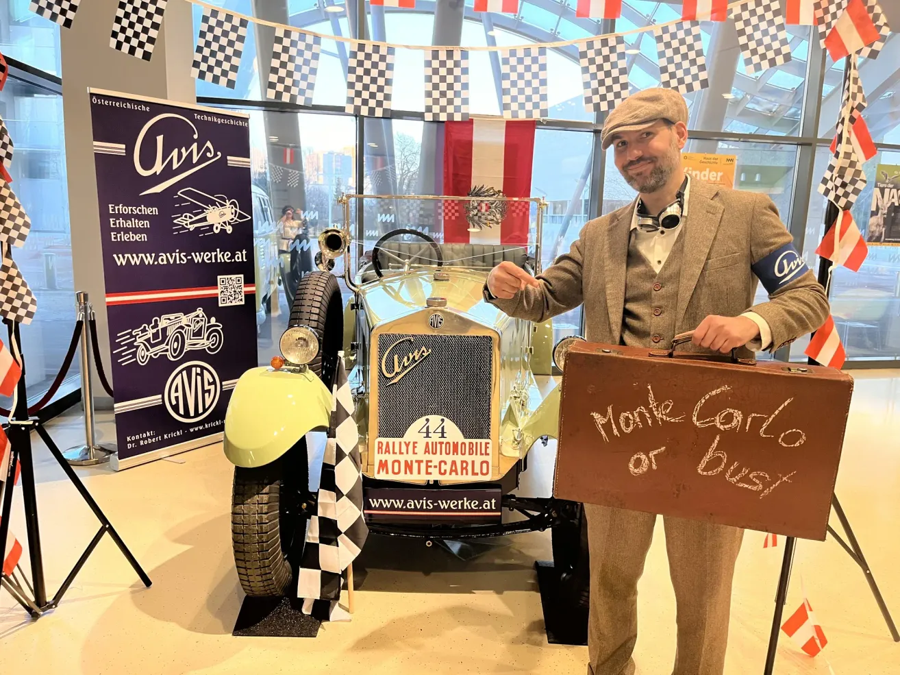 A man in a suit and hat stands next to a vintage car, holding a briefcase with the words 'Monte Carlo or Bust' written on it. The car is a rally automobile with the number 44 on its front. Behind him is a banner with the text 'AVIS' and a logo.