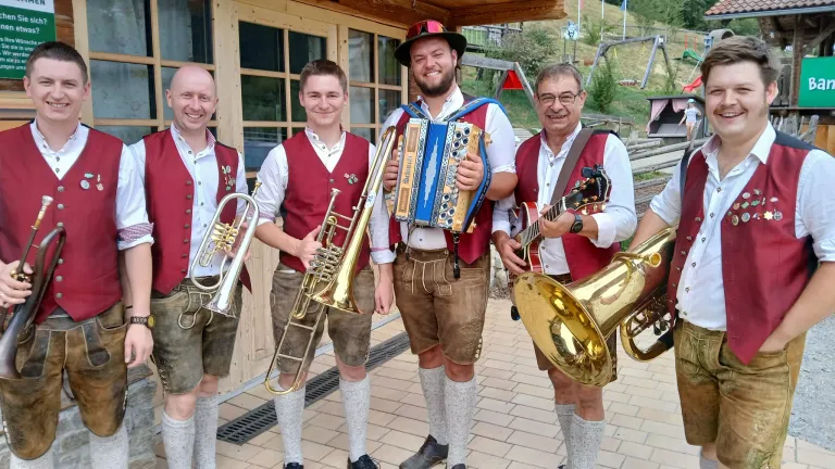 Four men dressed in traditional Bavarian attire, smiling and posing with musical instruments in front of a wooden building.