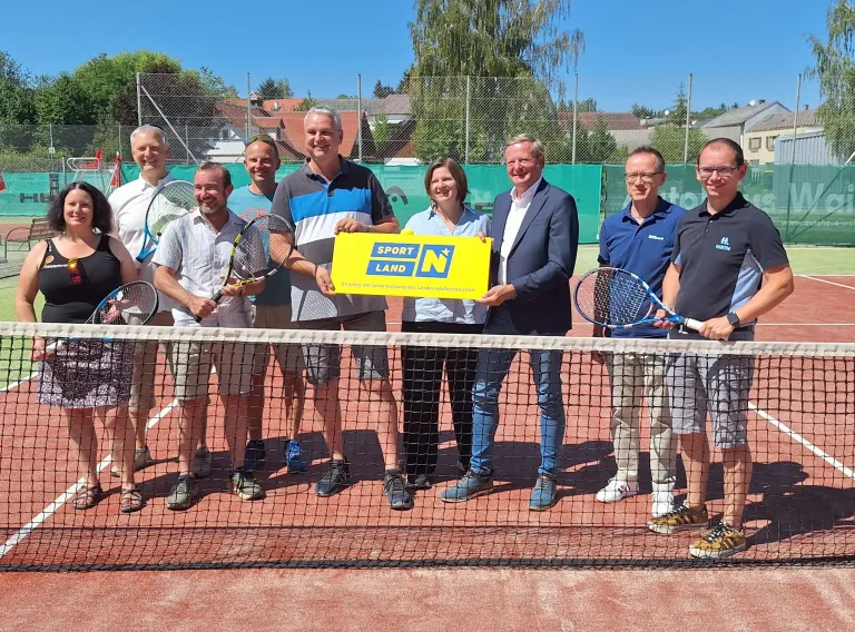 A group of people stand on a tennis court, holding tennis rackets, and posing for a photo with a yellow banner that reads 'SPORT LAND N'.