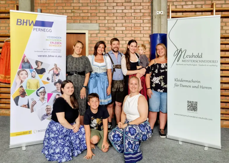 A group of people, including women in traditional Bavarian clothing and a man with a baby, pose for a photo in front of a brick wall and two banners.