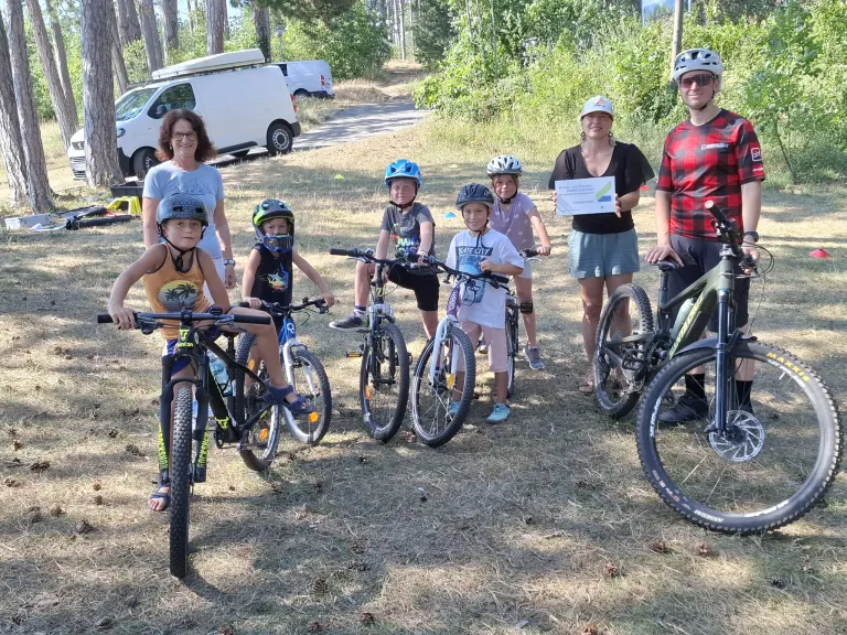 A group of children and adults are standing in a field with bicycles, some wearing helmets. A woman holds a certificate.