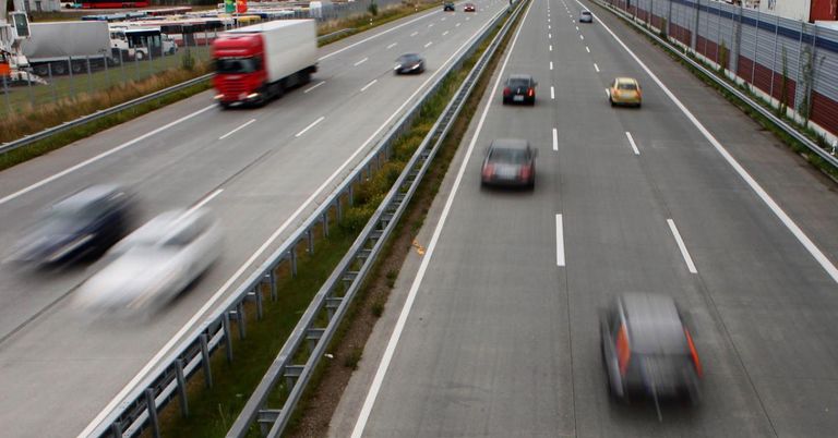 Eine Autobahn mit mehreren Fahrspuren, auf der ein roter Lkw und mehrere Autos in die gleiche Richtung fahren. Die Straße hat weiße Markierungen und eine Metallbarriere an der Seite.