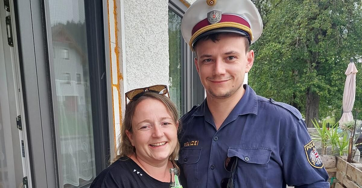 A smiling woman in a black shirt stands next to a smiling man in a blue police uniform, both posing for a photo in front of a building.