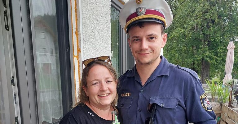 A smiling woman in a black shirt stands next to a smiling man in a blue police uniform, both posing for a photo in front of a building.