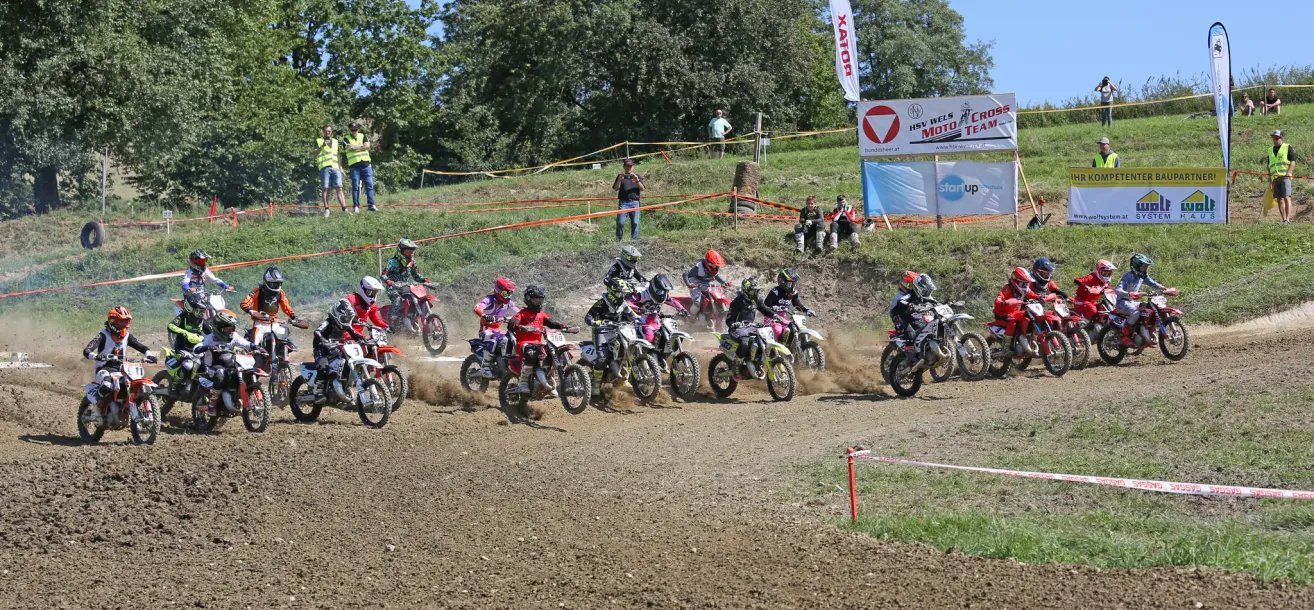 Eine Gruppe junger Rennfahrer nimmt an einem Motocross-Rennen auf einer Schlammpiste teil, wobei Zuschauer von den Seitenlinien zuschauen.