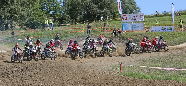 Eine Gruppe junger Rennfahrer nimmt an einem Motocross-Rennen auf einer Schlammpiste teil, wobei Zuschauer von den Seitenlinien zuschauen.