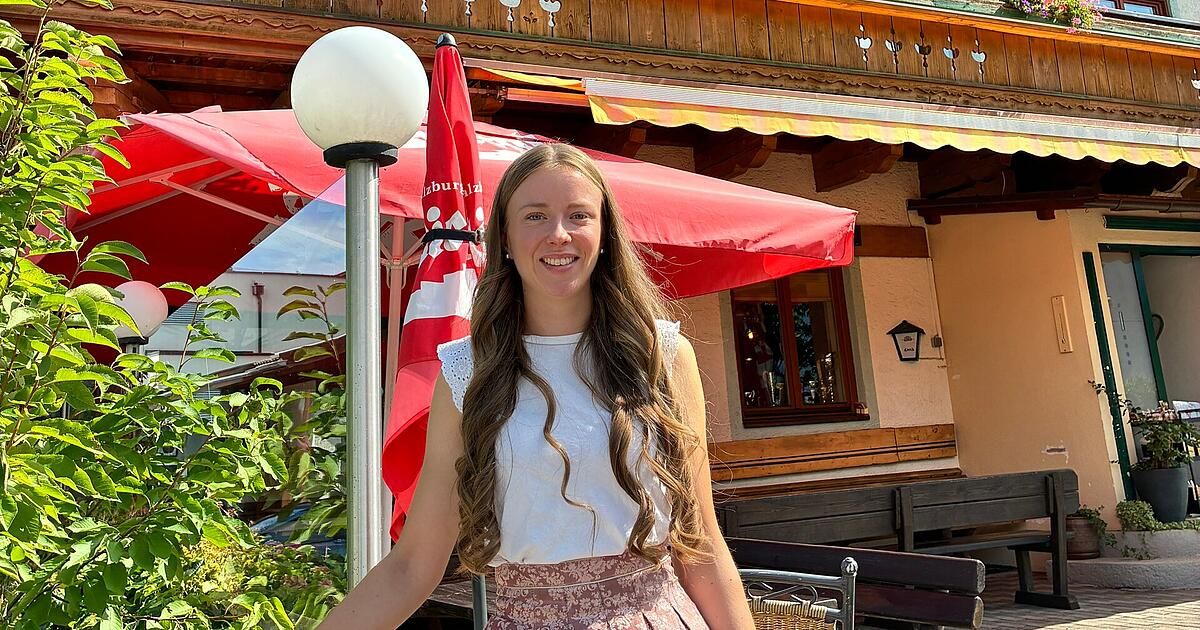 A woman with long curly hair stands outside a restaurant, smiling and posing for a photo. She is wearing a white blouse and a floral skirt. Behind her, there is a red umbrella and a lamp post. The restaurant has wooden benches and a window with a view of a car.