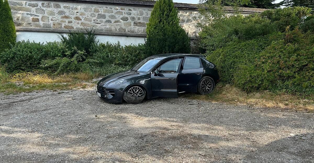 A black Porsche SUV with a flat tire is parked on a gravel driveway next to a stone wall.