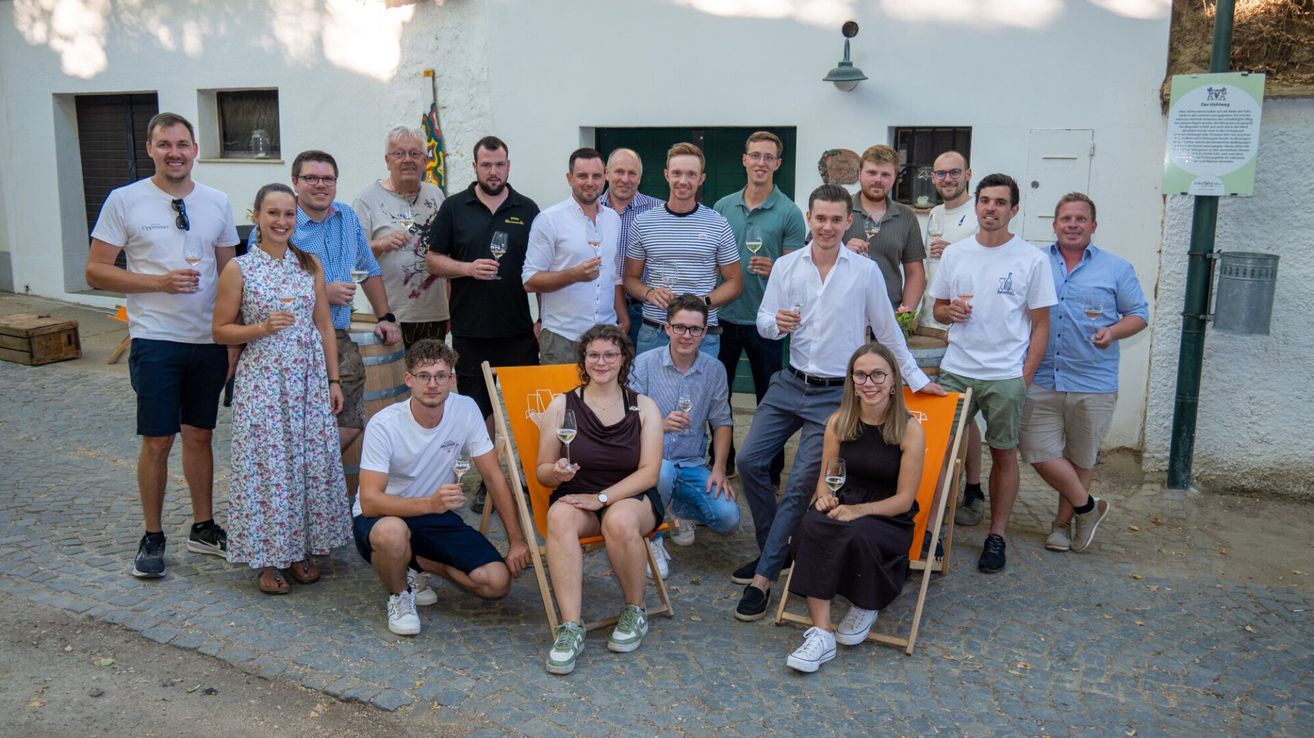 Eine Gruppe von Menschen in Freizeitkleidung, die Weingläser halten, posiert für ein Foto vor einem weißen Gebäude. Einige sitzen auf Stühlen, während andere stehen.