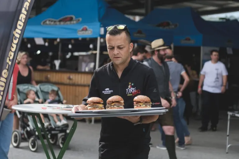 A man wearing a black shirt with a logo on it is carrying a tray with three burgers at an outdoor event.