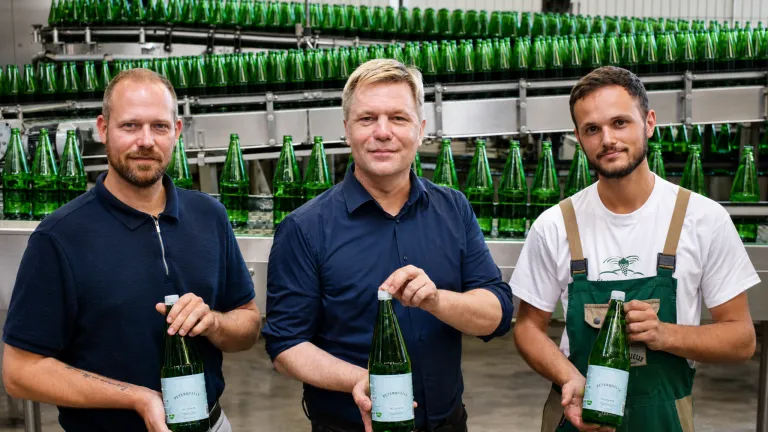 Three men stand in a factory, holding green bottles. The man in the middle smiles and holds a bottle with a white label. Behind them is a conveyor belt with more green bottles.
