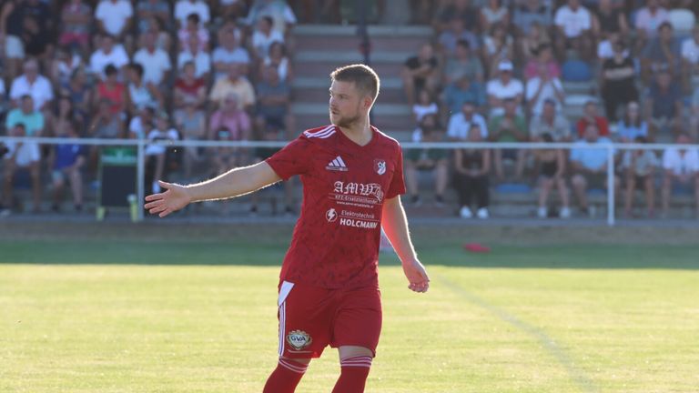 A soccer player in a red uniform with white stripes is gesturing with his arms during a match.