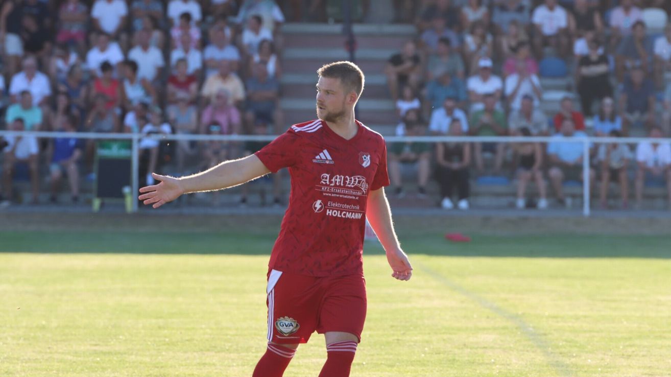 A soccer player in a red uniform with white stripes is gesturing with his arms during a match.