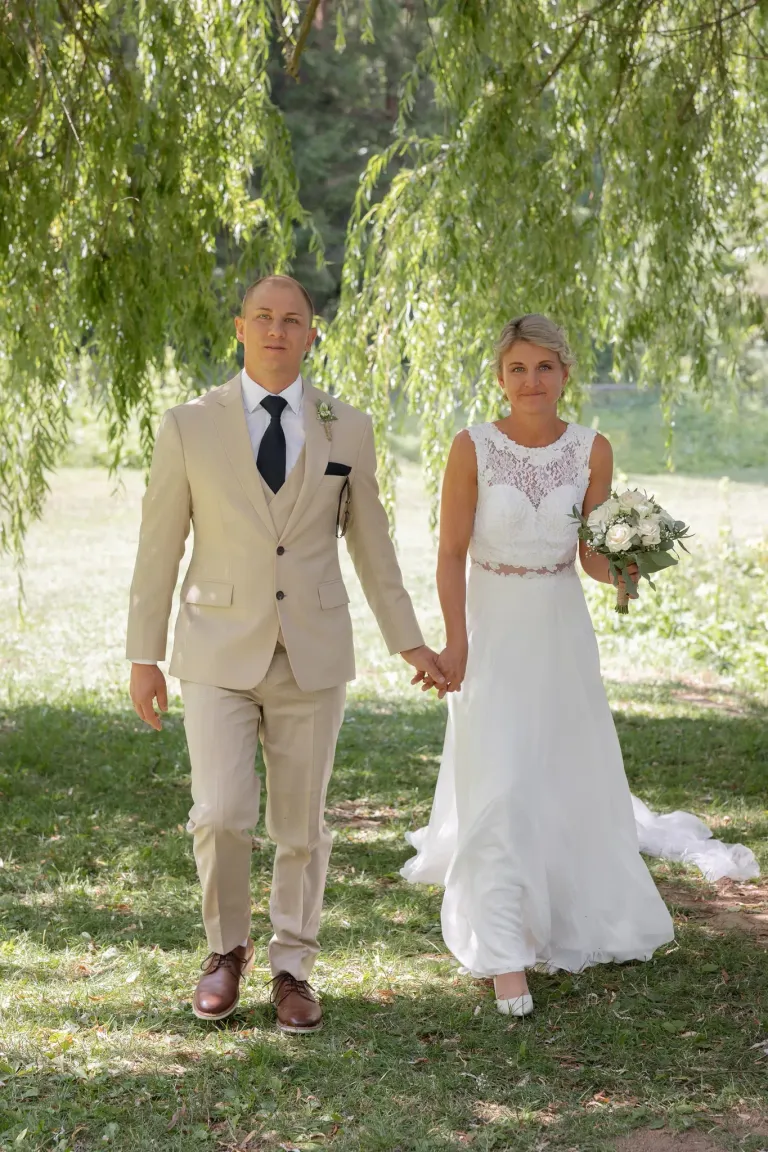 A man and woman in wedding attire are holding hands and walking in a grassy area under a willow tree.