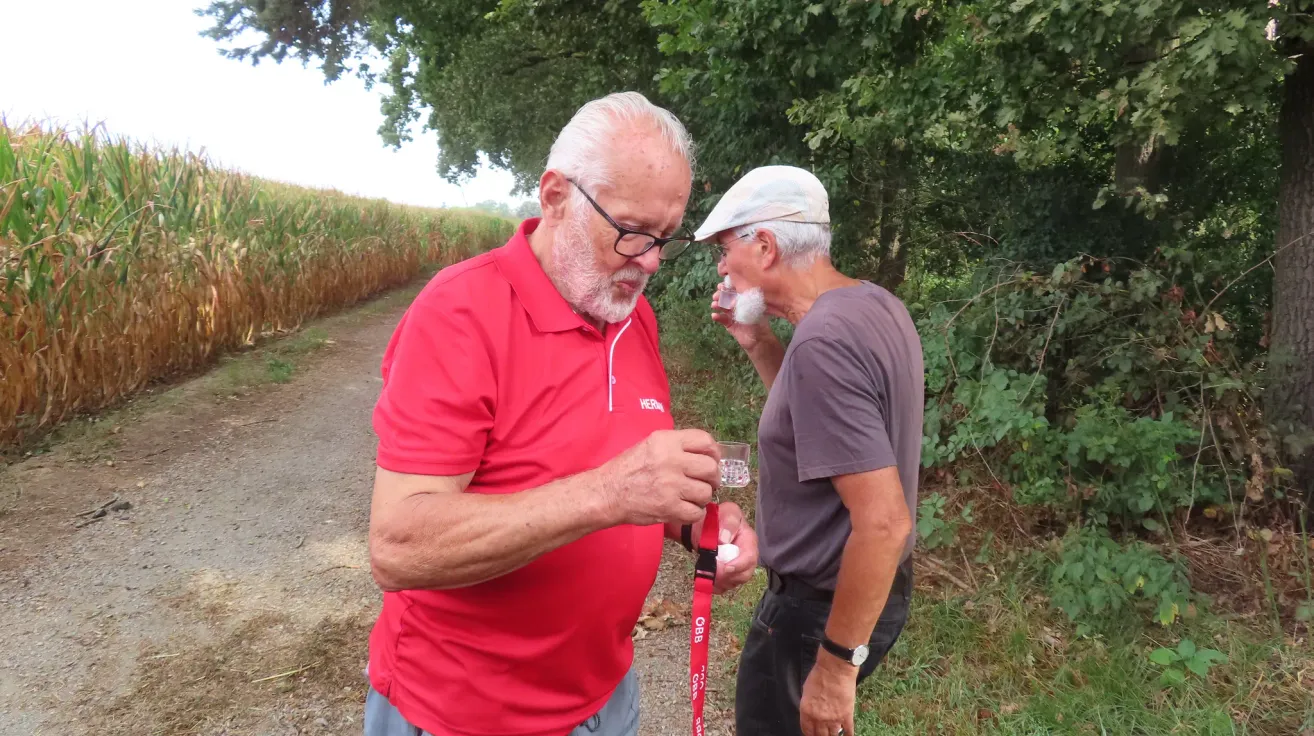 Two older men stand on a path in a forest, one drinking from a glass while the other holds a glass and a red lanyard.