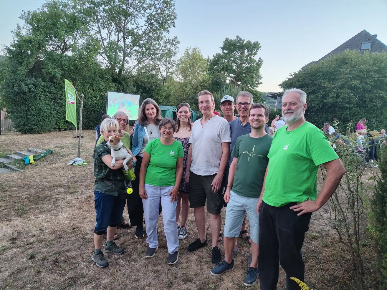 A group of people, some in green shirts, are standing together in an outdoor setting. A woman holds a dog, and a man in a green shirt has his hands on his hips. Trees and a green flag are in the background.