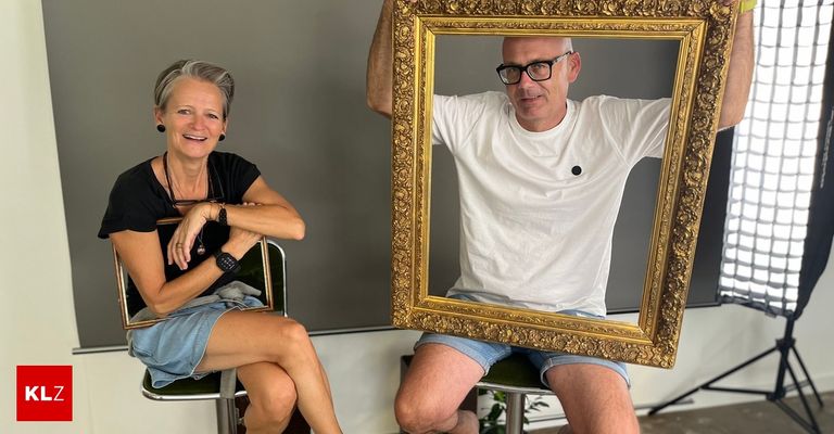 Ein Mann und eine Frau sitzen auf Stühlen, lächeln und halten einen großen, verzierten goldenen Bilderrahmen. Der Mann trägt eine Brille und ein weißes T-Shirt, während die Frau ein schwarzes Shirt und Jeansshorts trägt.