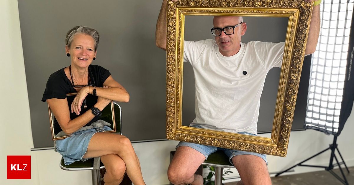 Ein Mann und eine Frau sitzen auf Stühlen, lächeln und halten einen großen, verzierten goldenen Bilderrahmen. Der Mann trägt eine Brille und ein weißes T-Shirt, während die Frau ein schwarzes Shirt und Jeansshorts trägt.