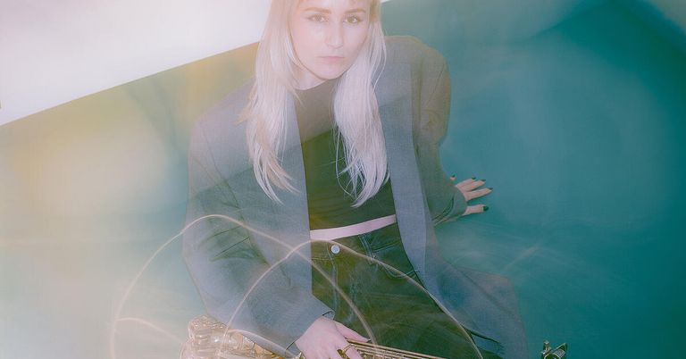 A woman with long blonde hair wearing a black crop top and gray blazer is sitting on a couch, holding a gold-colored object. The background is blurry.
